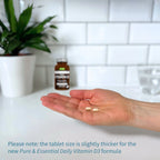 Hand holding two tablets with a bottle of Vitamin D3 in the background on a white surface.