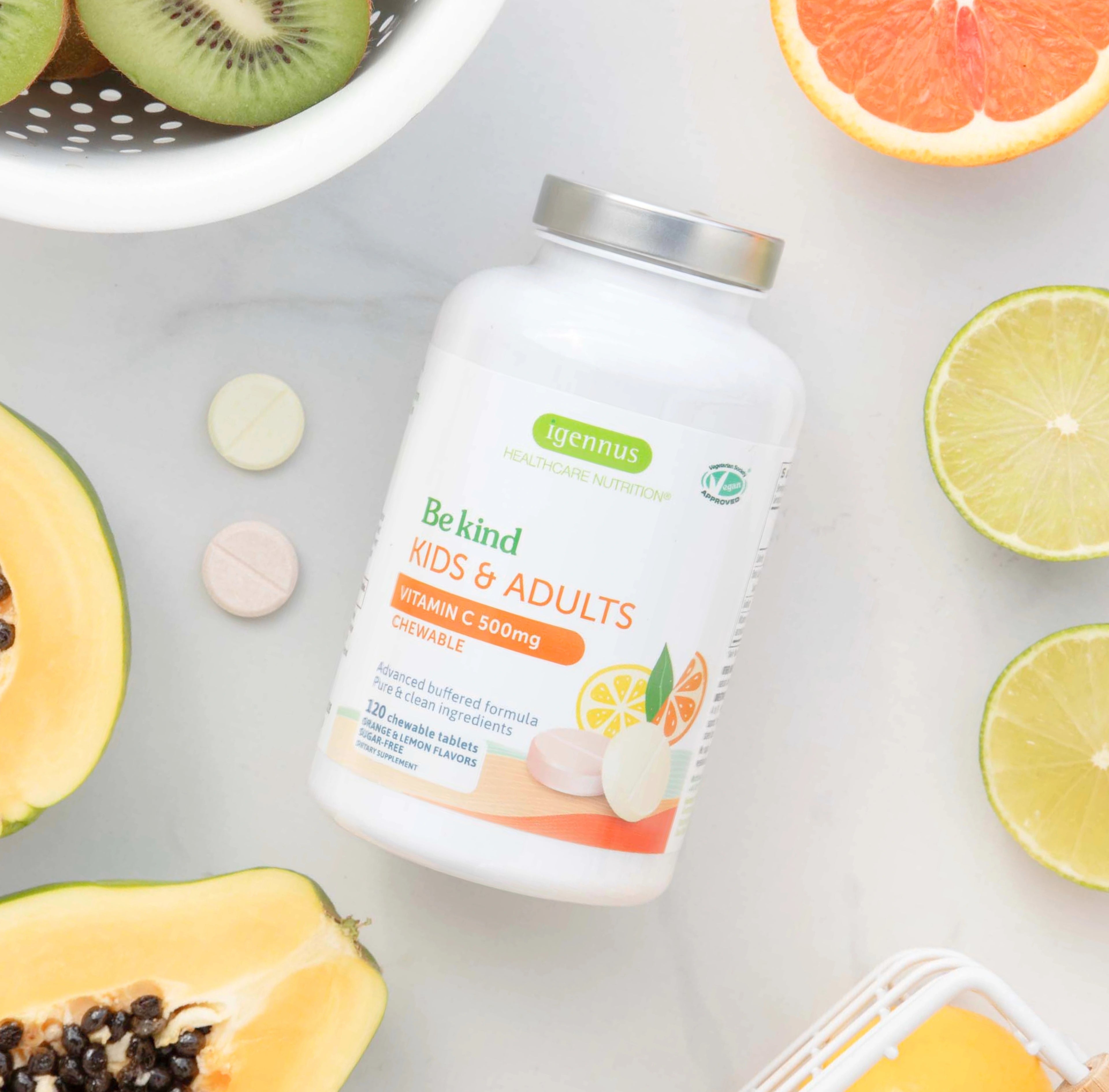 Vitamin supplement bottle labeled 'Be kind KIDS & ADULTS' surrounded by fruits on a light surface.