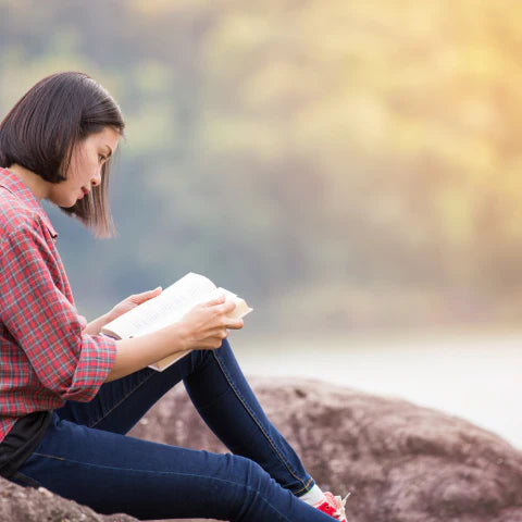 Woman reading a book outdoors on a rock with a blurred natural background