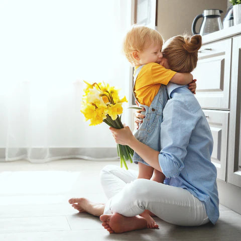 Woman sitting on the floor with a child, holding yellow flowers in a bright room.
