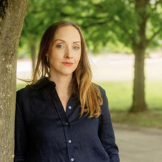 Woman standing outdoors in a park-like setting with trees and greenery.