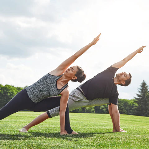 Two people practicing yoga on a grassy field with trees in the background