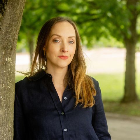 Woman standing outdoors under a tree with greenery in the background