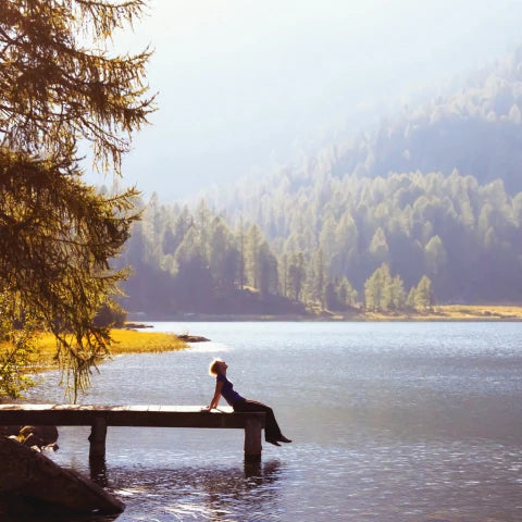 Person sitting on a wooden bench by a lake with trees and mountains in the background
