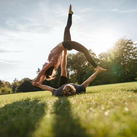 Two people practicing acrobatics in a park with trees and sunlight.