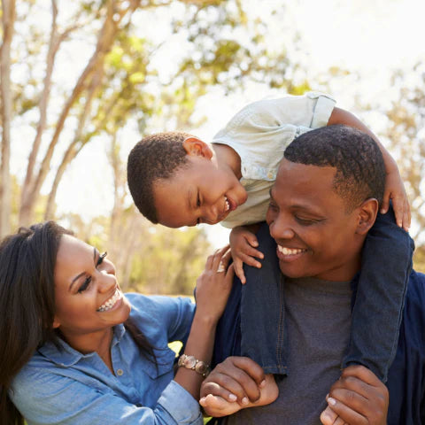 Family of three outdoors with a child on the father's shoulders, smiling.