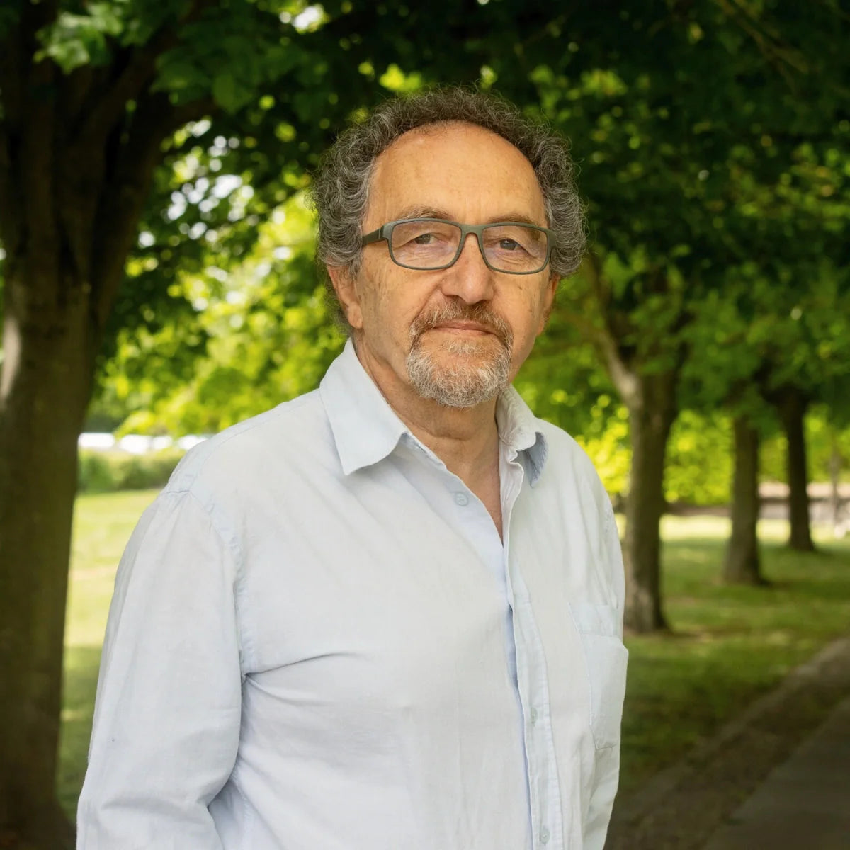 Man wearing glasses and a light shirt standing in a park with trees and greenery.