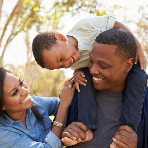 Family of three outdoors, with a father holding a child on his shoulders and a mother smiling.