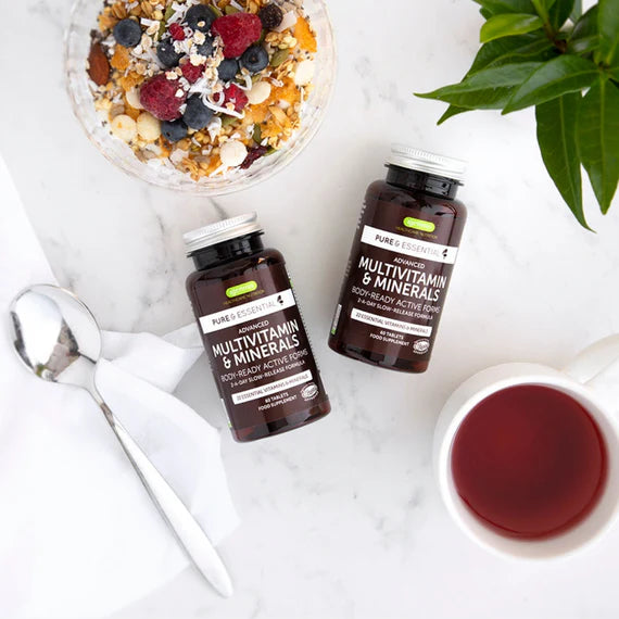 Two bottles of Pure &天然 multivitamin and mineral supplements on a white surface with a bowl of fruit, spoon, and mug.