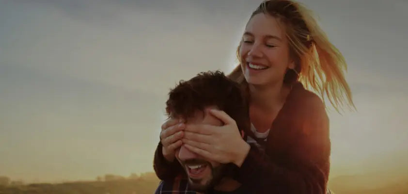 Woman playfully covering a man's eyes with her hands against a sunset sky.