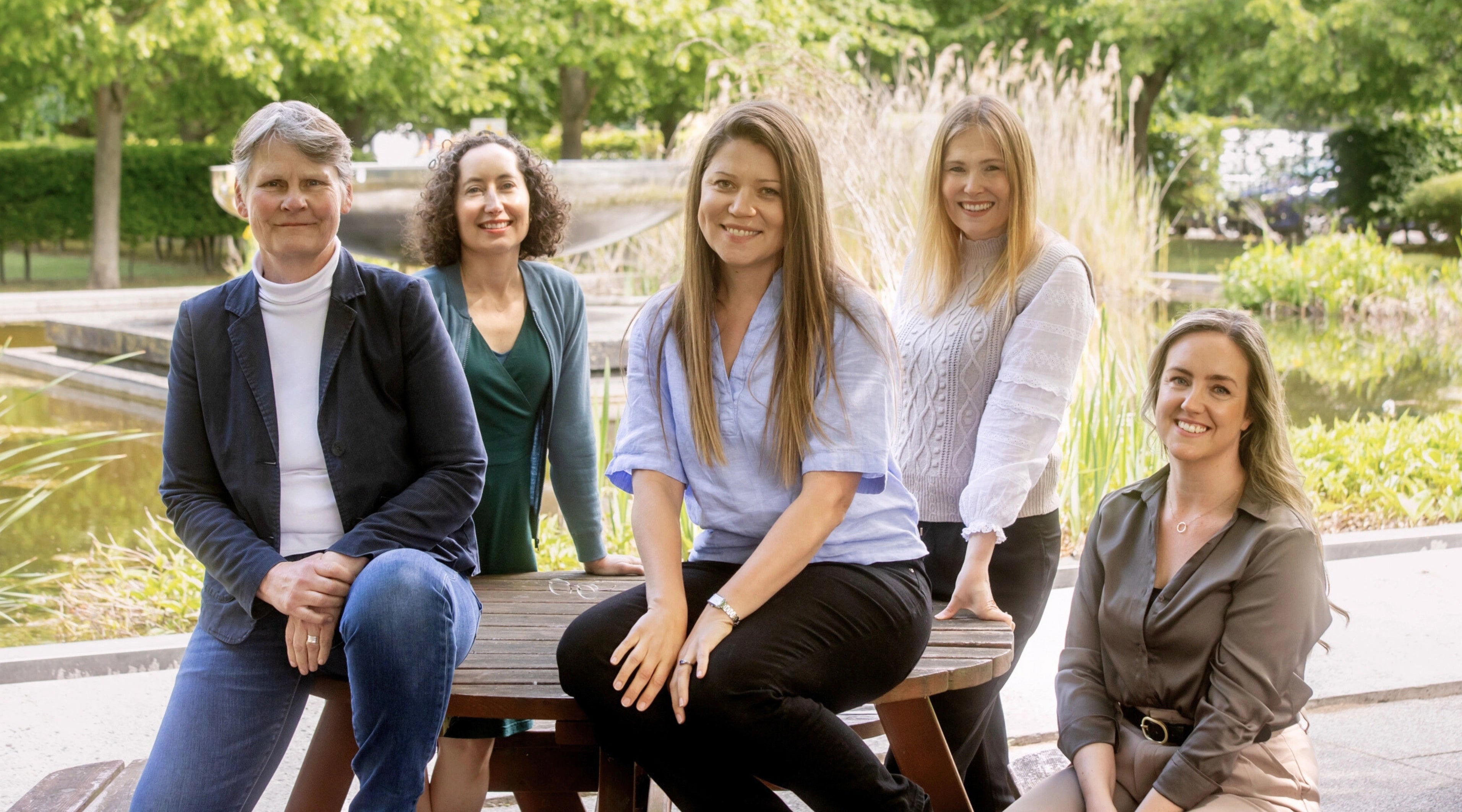 Five women sitting on a bench outdoors with trees and water in the background
